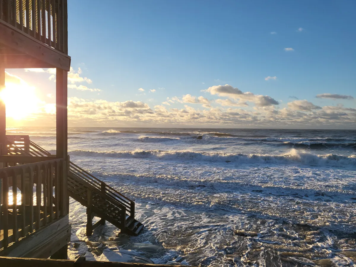 This image captures a spectacular sunset over the ocean, with gentle waves rolling onto the shore and wooden stairs leading down to the sand. The composition invites viewers to imagine a romantic stroll or quiet moment, bathed in the warm glow of dusk.
