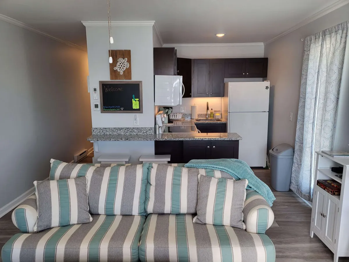 An open-concept living and kitchen area featuring a striped couch, granite-topped breakfast bar, and modern kitchen appliances. A cheerful 'welcome' sign and warm, homey décor tie the space together.