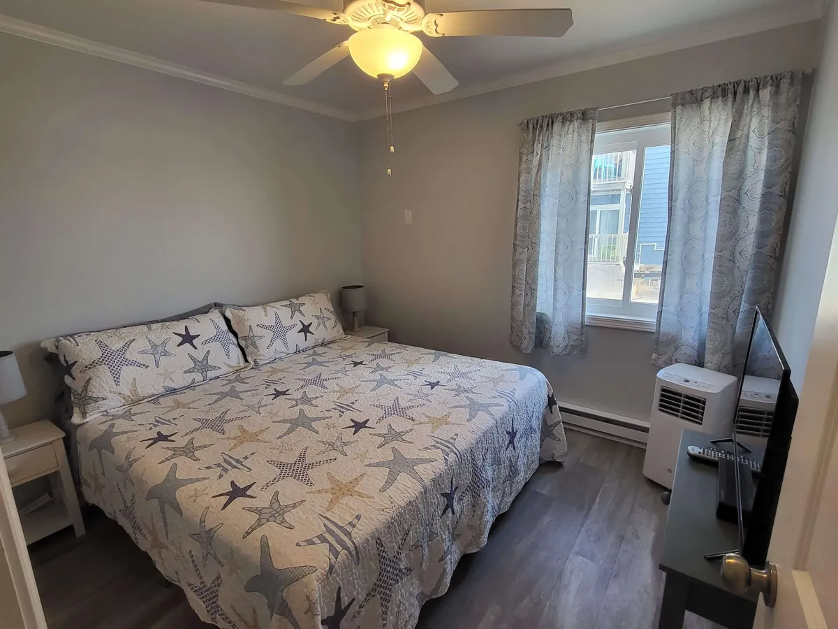 This image presents a bed adorned with a starfish-patterned comforter, accompanied by a nightstand with a lamp and a window dressed in sheer curtains. The atmosphere is calm and inviting, with natural light softly illuminating the space, evoking a sense of tranquility and comfort.