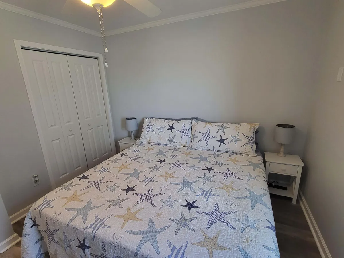A relaxing bedroom scene with a starfish-patterned quilt on a comfortable bed, flanked by nightstands with lamps and closet doors in the background. The space is neat, light-filled, and styled with modern coastal accents.