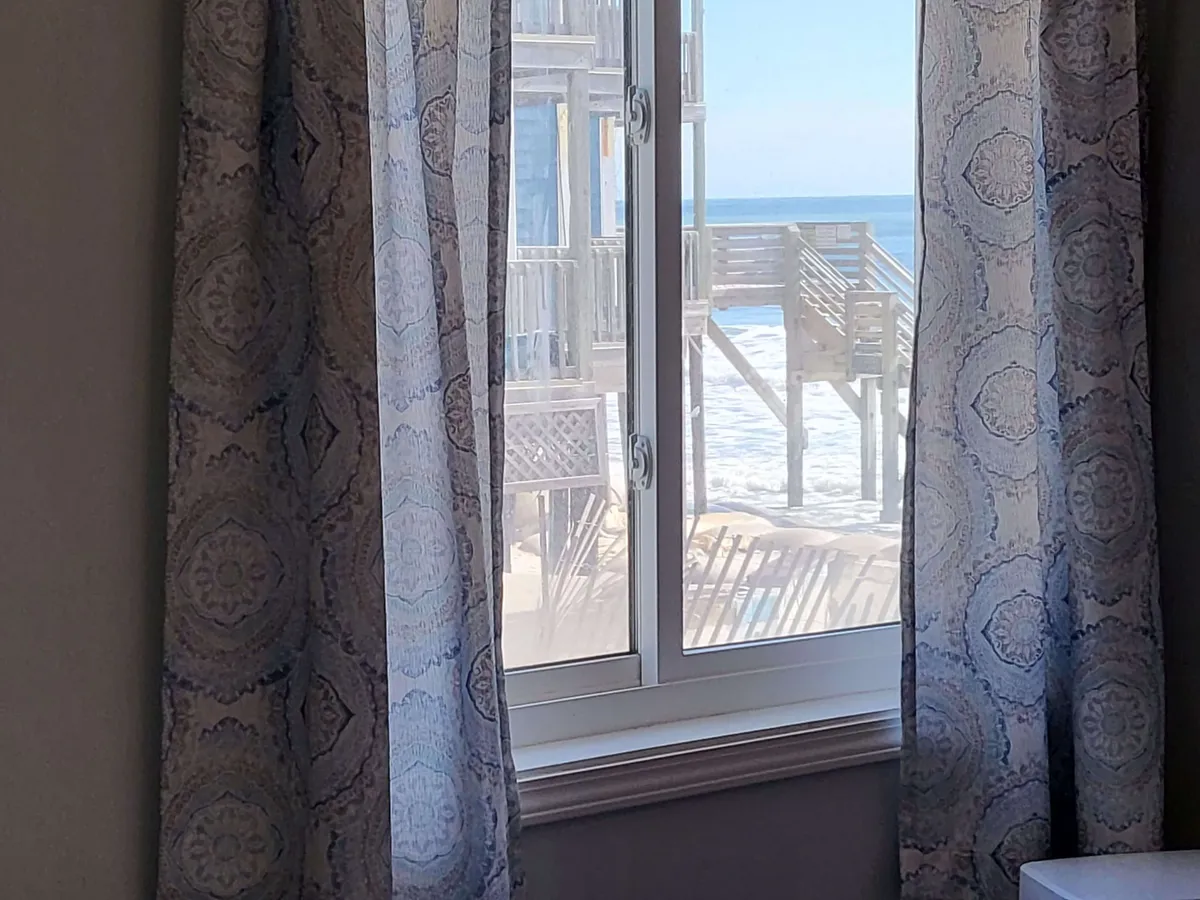 Interior view through a window with curtains, revealing a serene ocean vista and a deck outside. The space is quiet, ideal for reading or unwinding while gazing out at the water and feeling the ocean breeze.
