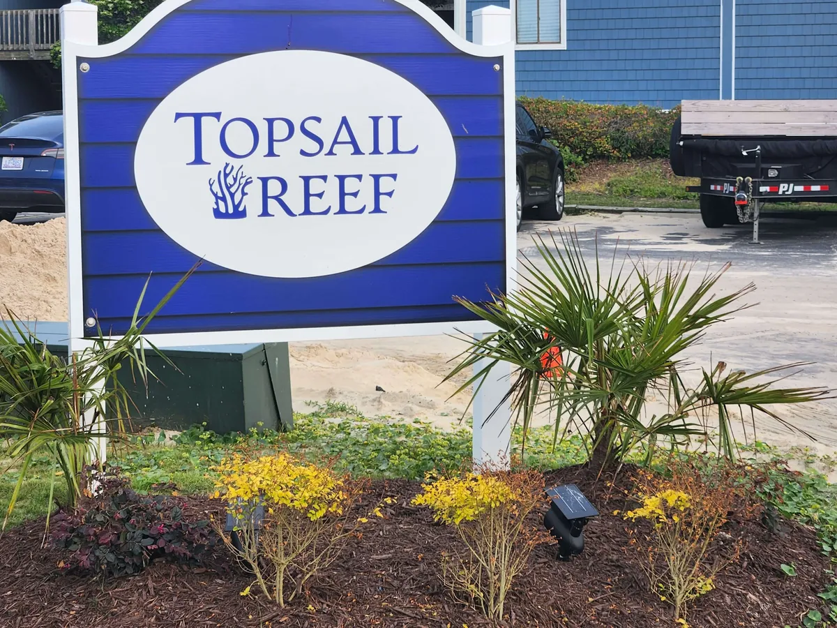The entrance to the property, marked by the 'Topsail Reef' sign, is decorated with lush landscaping and a background building, setting a welcoming tone for arrivals and departures.