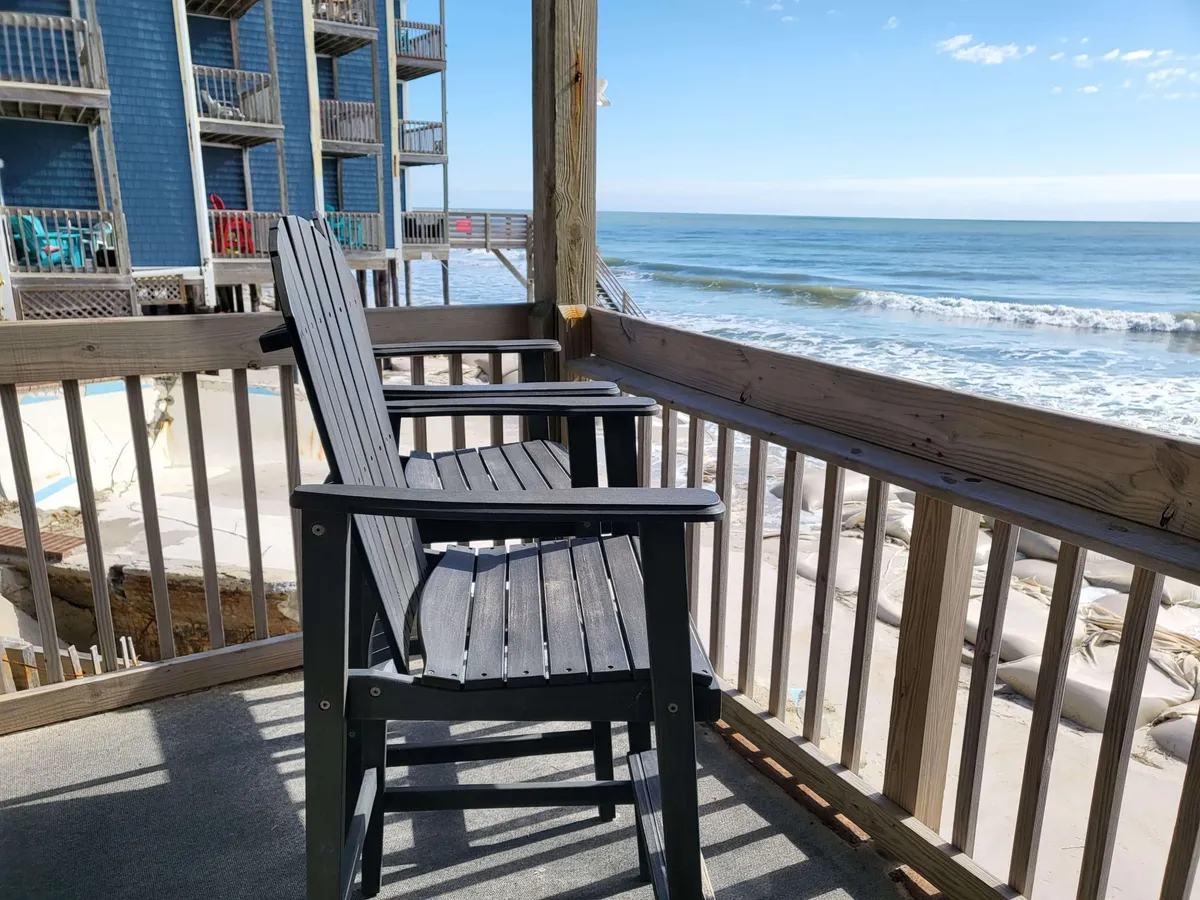 The image showcases a serene first-floor oceanfront balcony with wooden chairs overlooking the sparkling blue Atlantic and a wide, sandy North Topsail Beach under a clear sky. The setting evokes relaxation, easy beach access, and peaceful moments enjoying the sights and sounds of the ocean.
