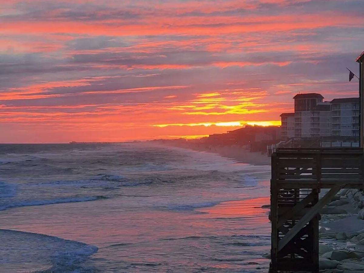A scenic sunset view with a long pier stretching into the ocean, waves lapping the shore, and beachgoers enjoying the evening. Buildings are visible in the background, evoking a sense of local amenities nearby.