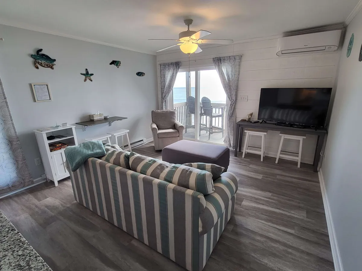 A relaxed interior scene featuring a striped sofa, TV, dining table, and an expansive ocean view through large windows. The space is casual and inviting, perfect for lounging or dining while enjoying the mesmerizing water just outside.