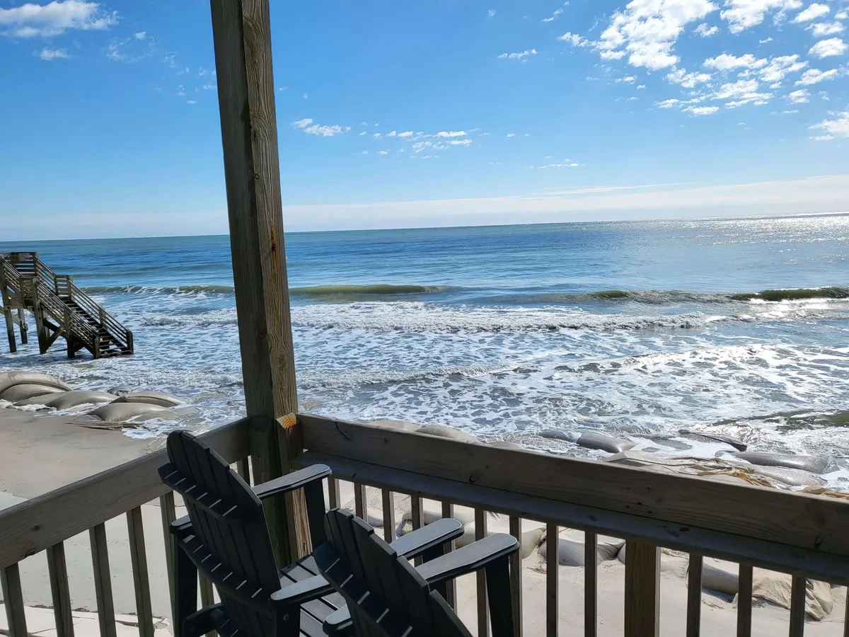 A serene oceanfront balcony set above the waves, featuring deck chairs, a wooden railing, and a staircase leading down to the sand. The scene evokes peaceful relaxation, with the ocean stretching out to the horizon and inviting sunrise or sunset moments.