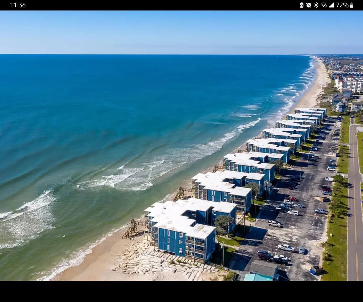 Beachfront apartments overlooking the ocean, with families and children enjoying the sand and waves. The parking lot and coastal road are visible, emphasizing easy access and a family-friendly atmosphere.