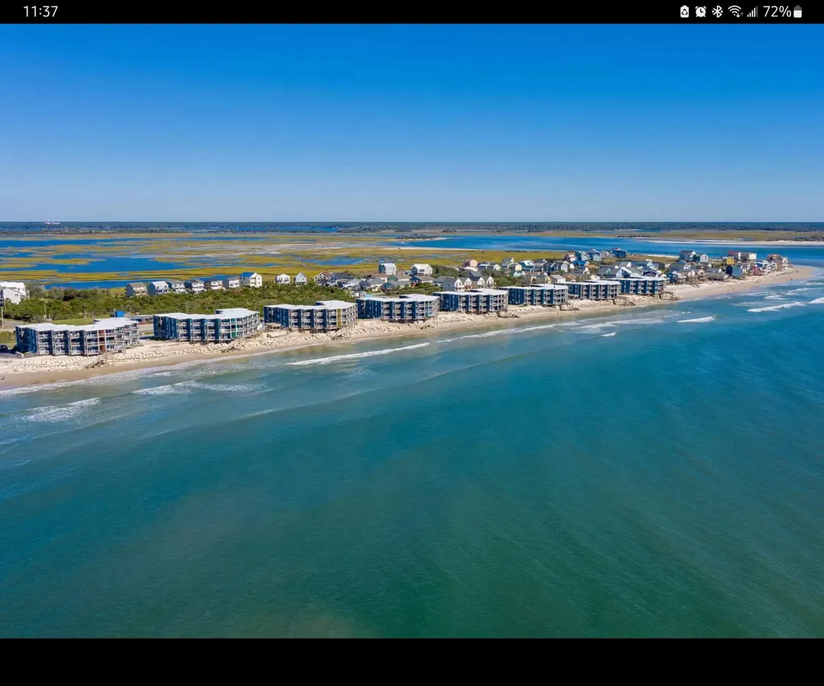 A wide view of beachfront properties with the ocean waves rolling in and marshlands visible in the distance. The sand looks inviting and the entire scene evokes the feel of immediate beach access, with the sun either rising or setting for a warm glow.